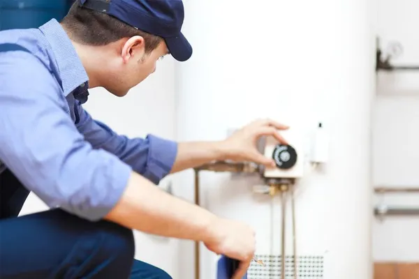 an HVAC technician in a blue shirt kneeling next to a water heater on a heating and plumbing service